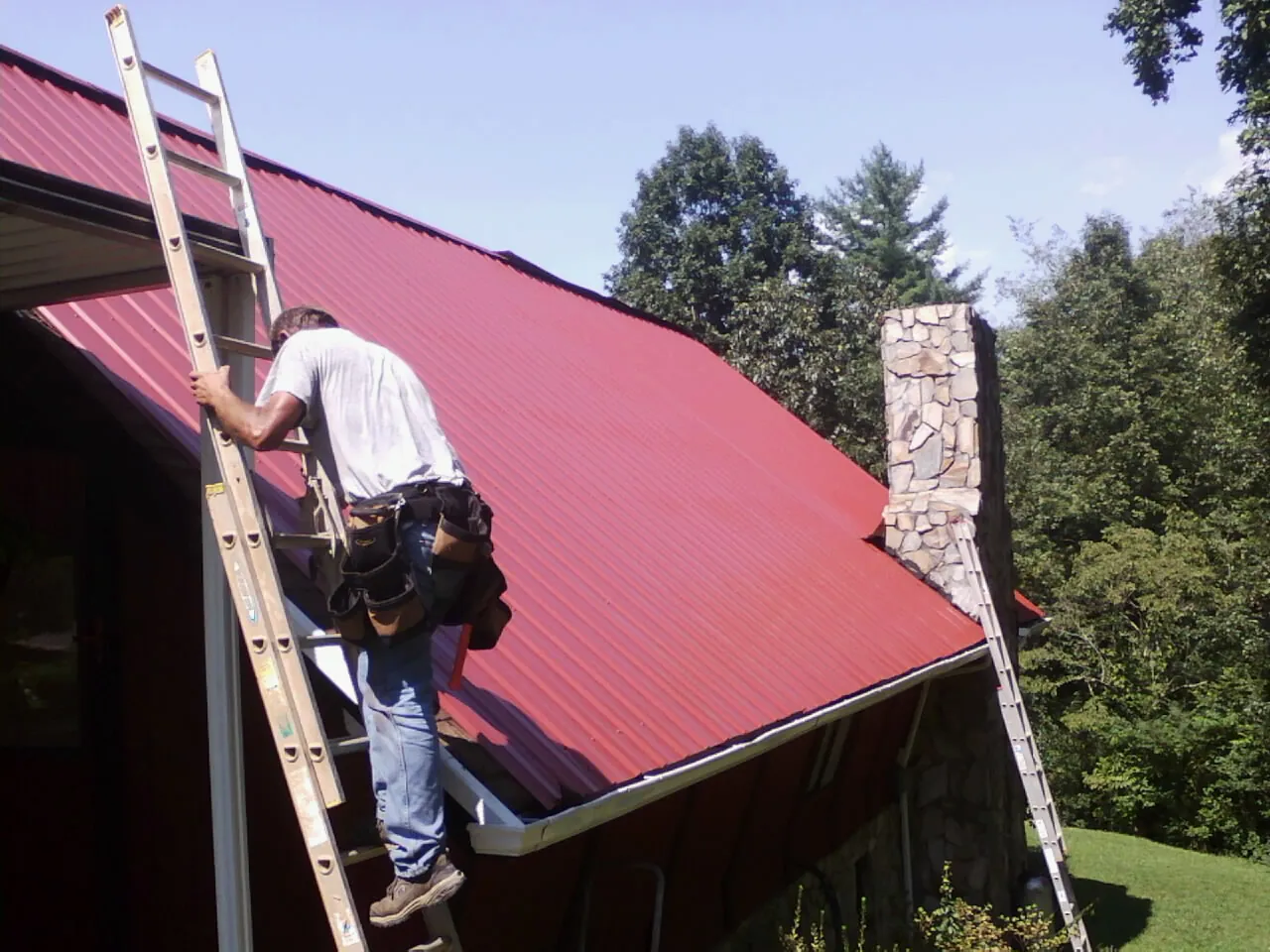 Roofer on a ladder beside a red metal roof and stone chimney — Soffit & Fascia Repair by Roofing Brothers in Melrose Park